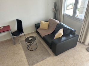 Ein Wohnzimmer mit einem schwarzen Ledersofa, einem roten Tisch, einem schwarzen Sessel, einem kleinen runden Tisch und einem Fenster mit Vorhängen.