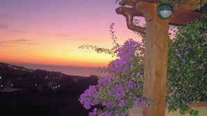 Une pergola en bois, ornée de fleurs violettes, d’où l’on peut admirer un coucher de soleil sur la silhouette d’une ville.