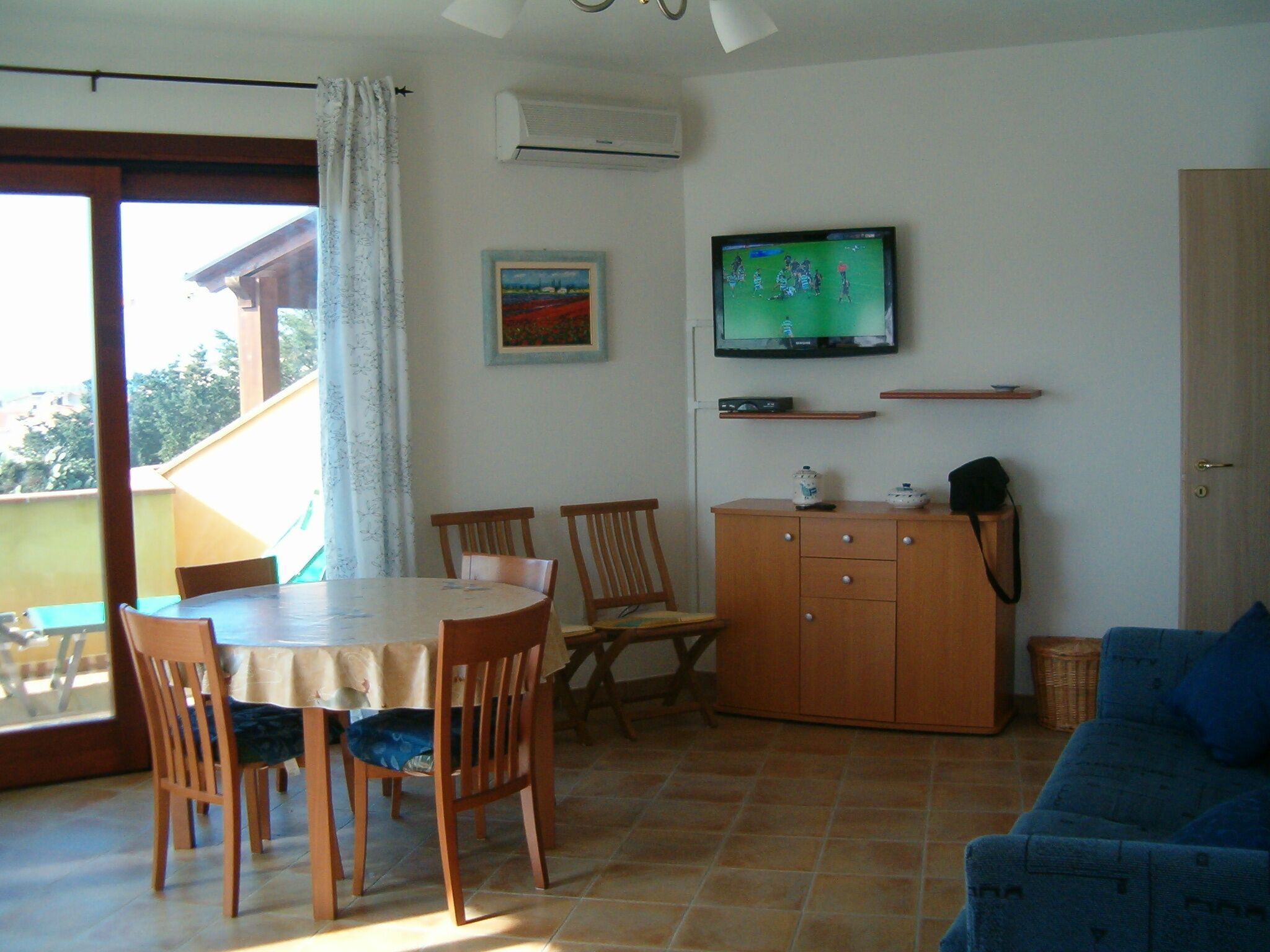 A dining area with a table and chairs, a television mounted on the wall, and a blue sofa.