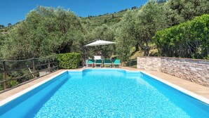 Ein rechteckiger Swimmingpool mit klarem, blauem Wasser, umgeben von einer Steinmauer und einem Holzzaun. Zwei Liegestühle mit grünen Kissen und ein weißer Sonnenschirm sind vorhanden.