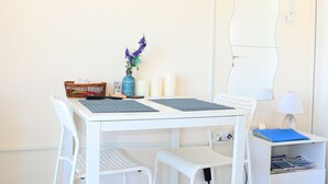 Une pièce moderne et minimaliste, avec un bureau blanc, deux chaises, un téléviseur à écran plat fixé au mur, une petite table d’appoint avec une lampe et un vase décoratif contenant des fleurs violettes.