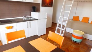 Eine kleine Küche mit Spüle, weißen Schränken und einer Kaffeemaschine. Ein Esstisch mit orangefarbenen Platzsets und Stühlen steht daneben. Im Hintergrund ist ein Etagenbett mit orangefarbenen Kissen zu sehen.
