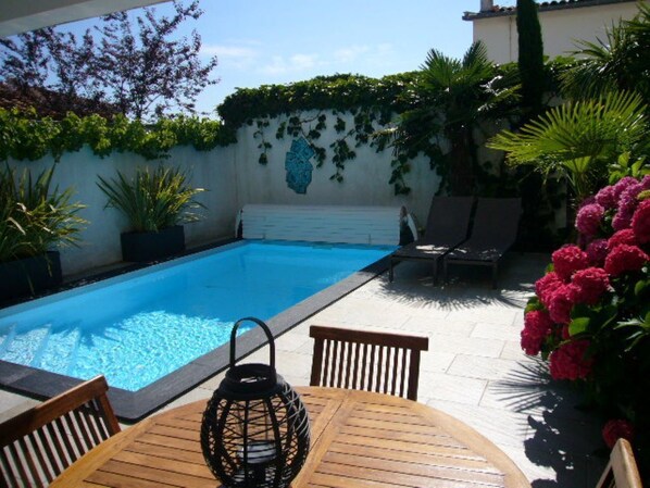 A swimming pool with a tiled deck, surrounded by potted plants and a wooden table with chairs.