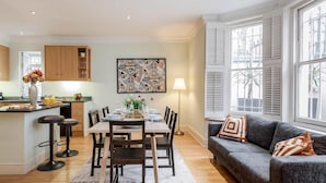 A modern kitchen with wooden cabinets, a dining area with a table set for four, and a grey sofa with patterned cushions. - Spacious 3 bed Kensington apartment with Patio (London)