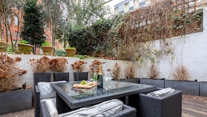 An outdoor patio with a glass-top table, wicker chairs, and a variety of plants and foliage. - Spacious 3 bed Kensington apartment with Patio (London)