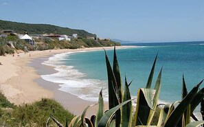 Uma praia com águas turquesa cristalinas, areal arenoso e alguns edifícios ao fundo.