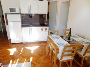 A compact kitchen with a microwave, sink, and cabinets, a dining table set with plates and glasses, and a bottle of wine.