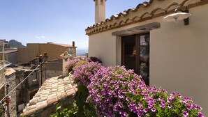 A building with a tiled roof, a chimney, and a window. There is a staircase leading to the entrance, and a row of pink flowers in front of the building. - "Domu Irde: from a ruin a dream" (Baunei)