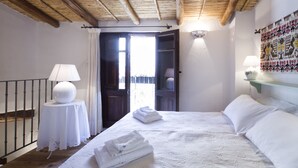 A bedroom with a bed, a bedside table with a lamp, a wall-mounted shelf, and a door leading to an outdoor area. - "Domu Irde: from a ruin a dream" (Baunei)