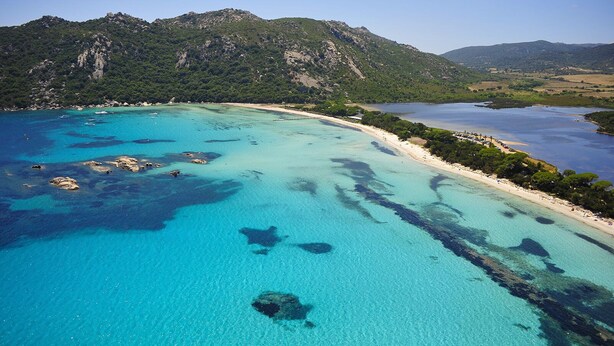 Luchtbeeld van een kustgebied met helder turquoise water, rotsachtige uitstulpingen en een bergachtige achtergrond.