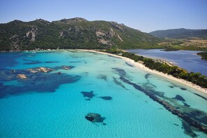 Vue aérienne d’une zone côtière avec des eaux turquoise limpides, des affleurements rocheux et un paysage montagneux en arrière-plan.