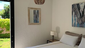 Een slaapkamer met een groot bed, een hanglamp, een lijst met een afbeelding, een nachtkastje met een lamp, en uitzicht op groen buiten.
