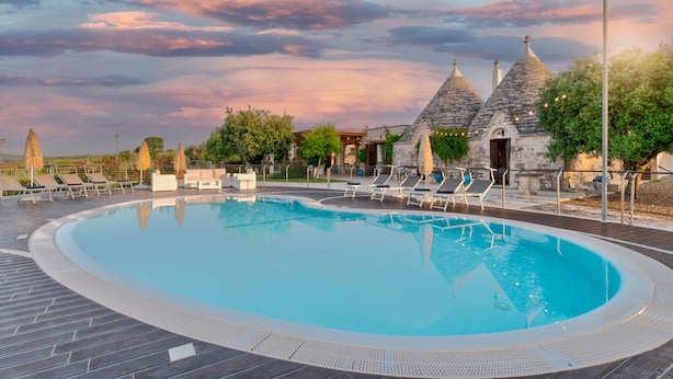 A swimming pool with lounge chairs, a stone building with conical roofs, and a sunset sky. - Luxury trullo, Private  Pool, Air con, Private Tennis Court, Pizza Oven (castellana grotte)