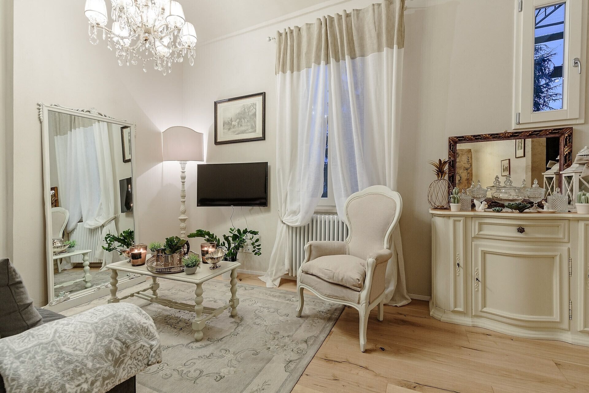 A living room with a chandelier, a television, a white armchair, a coffee table with plants, and a cabinet with decorative items.