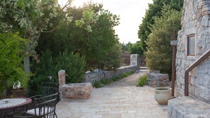 Um caminho de pedra serpenteia por um jardim, com uma mesa e cadeiras, rodeado por vegetação e muros de pedra.