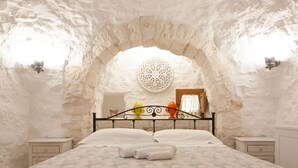 Quarto com parede de pedra, luminária redonda no teto, cama com almofadas e mesas de cabeceira.
