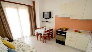 A compact hotel room with a bed, a dining table with chairs, a TV, a kitchen area with a stove and oven, and a window with curtains.