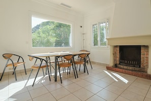 A dining area with a table and chairs, large windows with a view of greenery, and a fireplace.