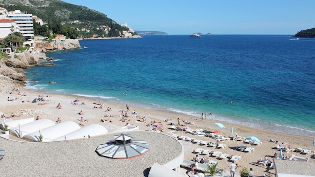 A beach with sun loungers and umbrellas, a clear blue sea, and buildings in the background.