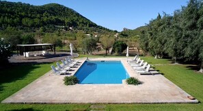 Piscina retangular com deck ao redor, cadeiras de sol e um espaço coberto para sentar.