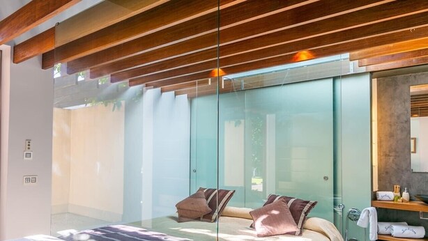 A modern bedroom with a glass partition, a bed with pillows, a wooden ceiling, and a bathroom with a sink and towel rack.