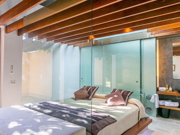 A modern bedroom with a glass partition, a bed with pillows, a wooden ceiling, and a bathroom with a sink and towel rack.