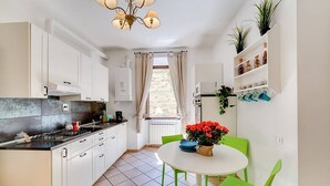 Une cuisine compacte avec des meubles blancs, une cuisinière à gaz et une fenêtre avec des rideaux. On y trouve un coin repas avec une table blanche et des chaises vertes, ainsi qu’une étagère contenant des ustensiles de cuisine et des plantes.