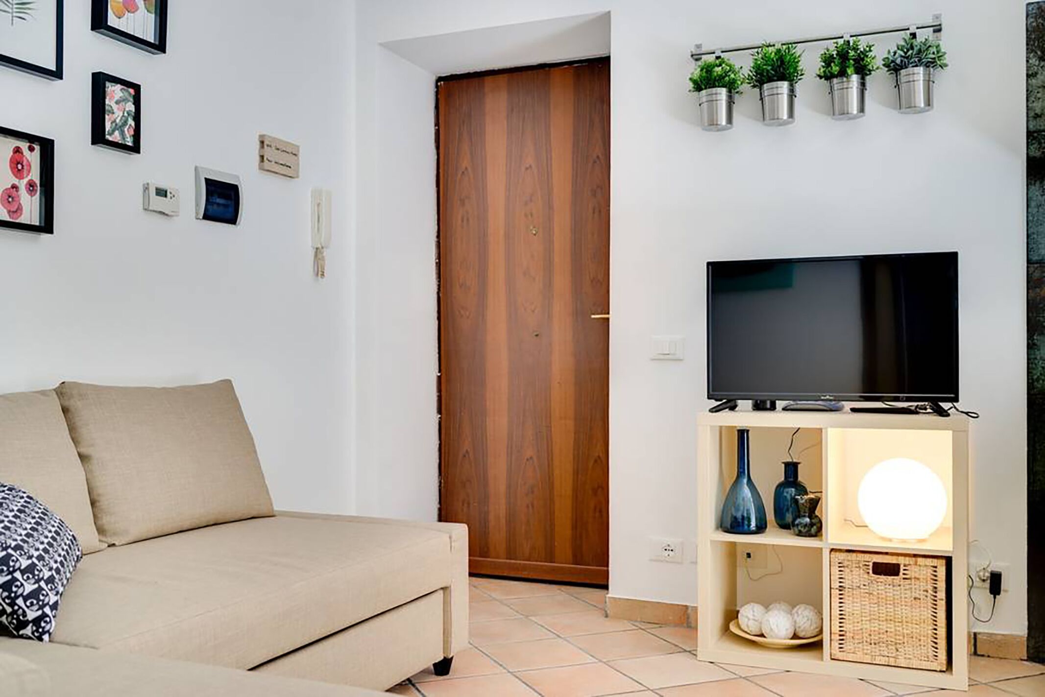 A living room with a beige sofa, a flat-screen TV on a wooden stand, a wall-mounted shelf with plants, and a door in the background.