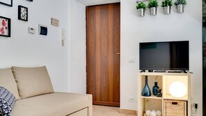 Un salon avec un canapé beige, un téléviseur à écran plat posé sur un meuble en bois, une étagère murale ornée de plantes et une porte visible au fond.