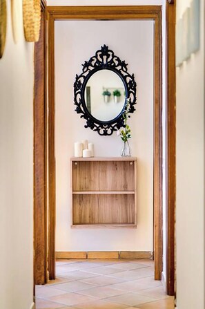 Enter a serene space with a hallway leading to a cozy room, featuring a door, mirror, and a shelf with candles and greenery.