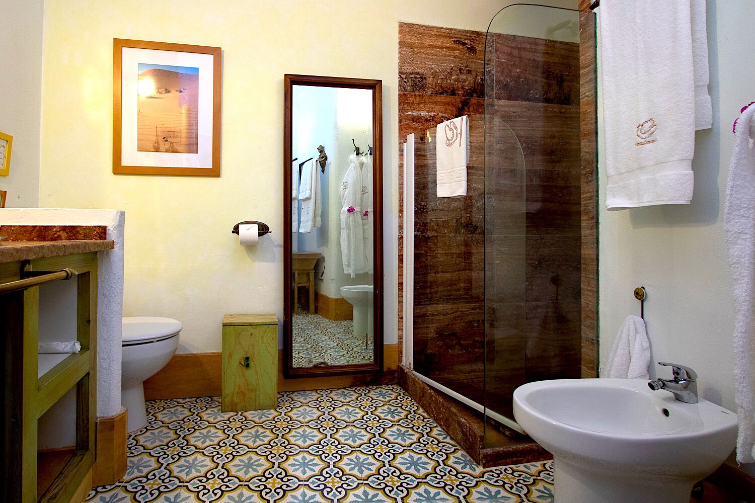 Une salle de bain avec des toilettes, un lavabo et une douche. On y trouve un miroir, un porte-serviettes et un tableau encadré au mur.