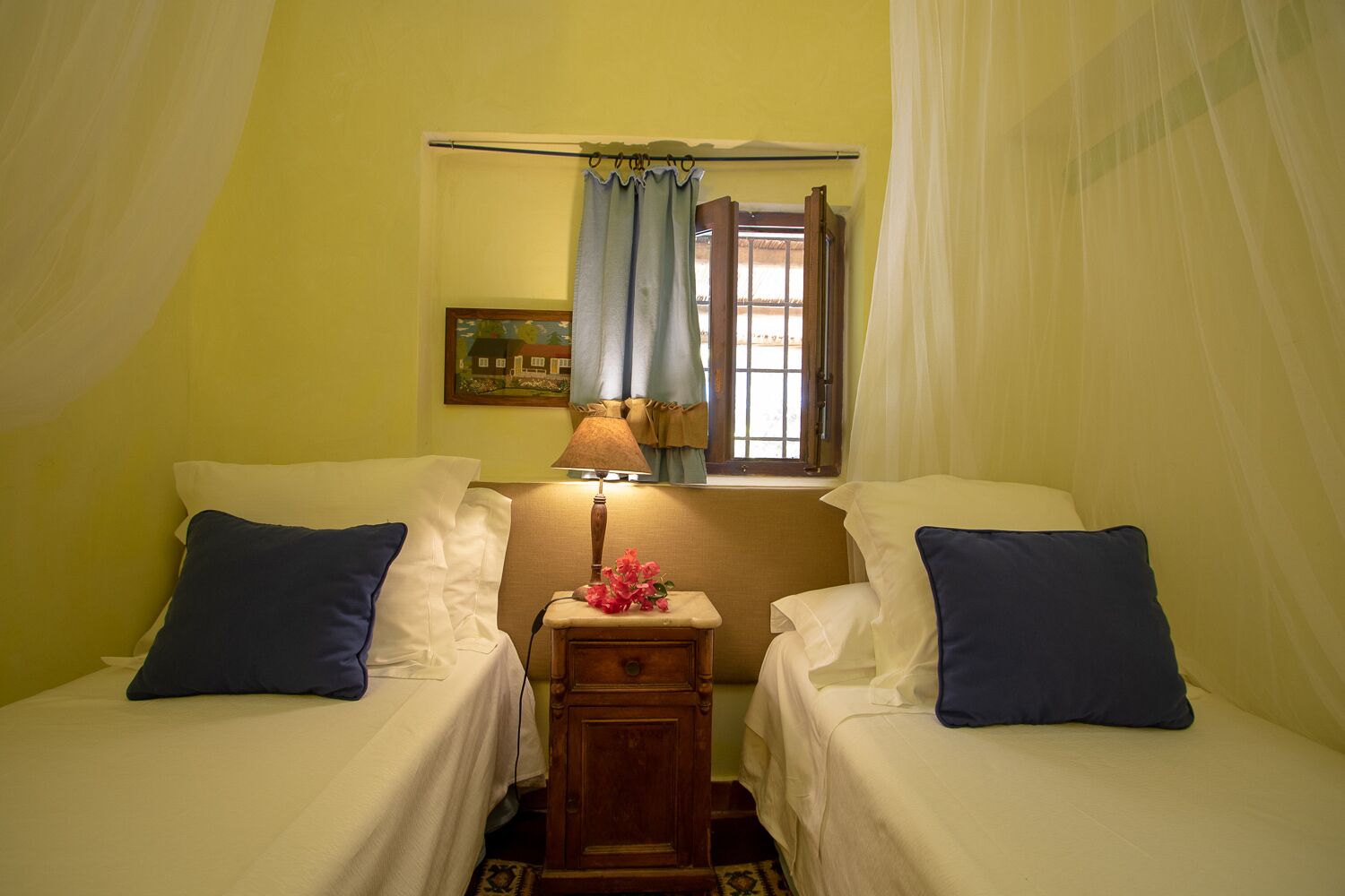 A room with two single beds, a nightstand with a lamp, a painting on the wall, and a window with curtains.