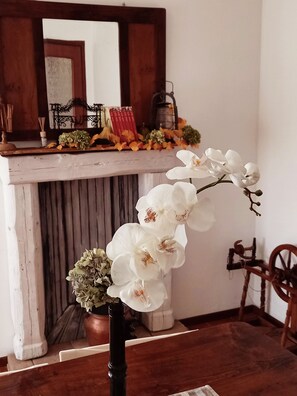 Une pièce avec une table en bois, une orchidée en pot et une cheminée surmontée d’une étagère décorée de livres et de fleurs.
