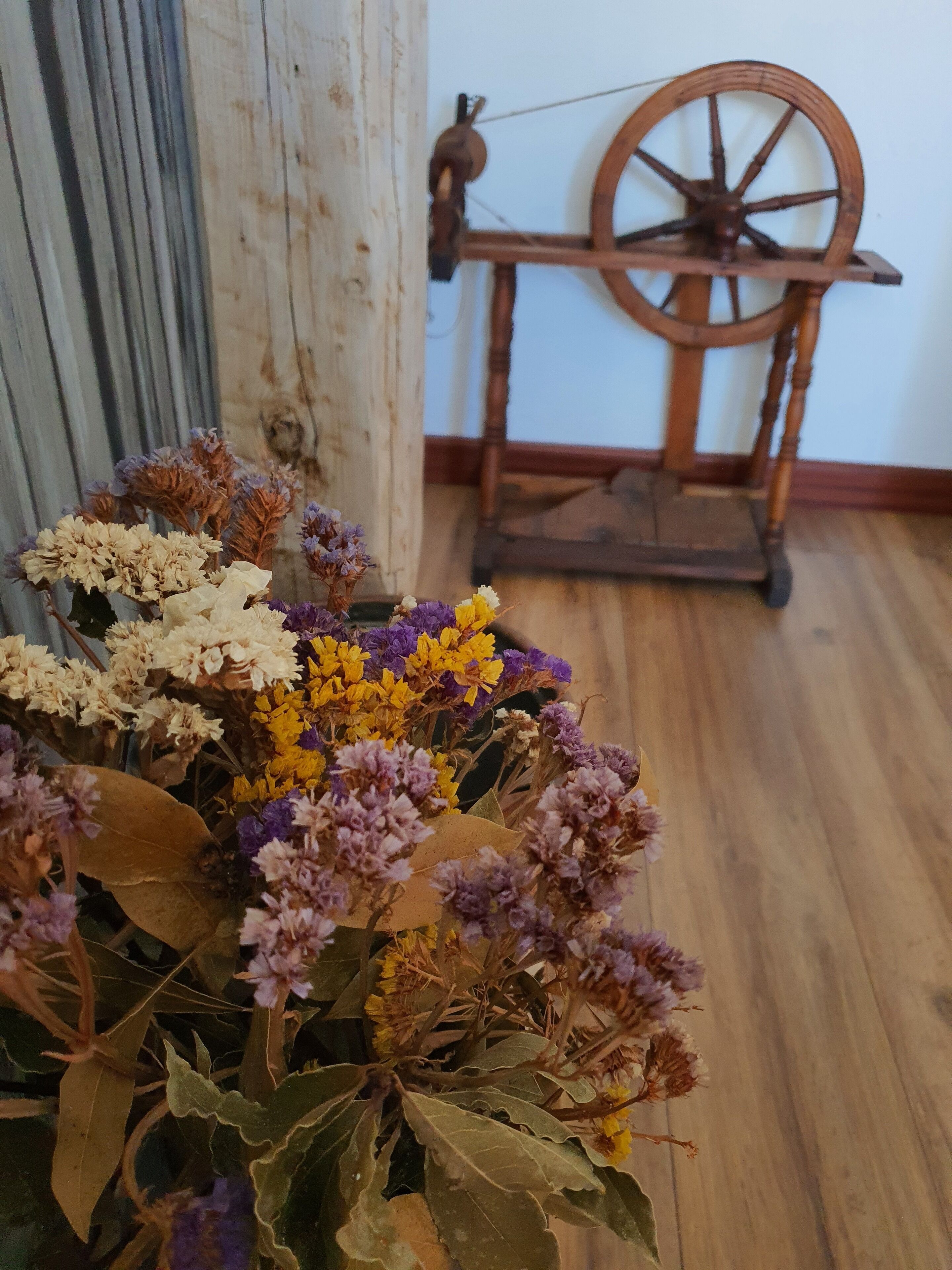 A room with a wooden floor, a spinning wheel, and a bouquet of dried flowers in the foreground.