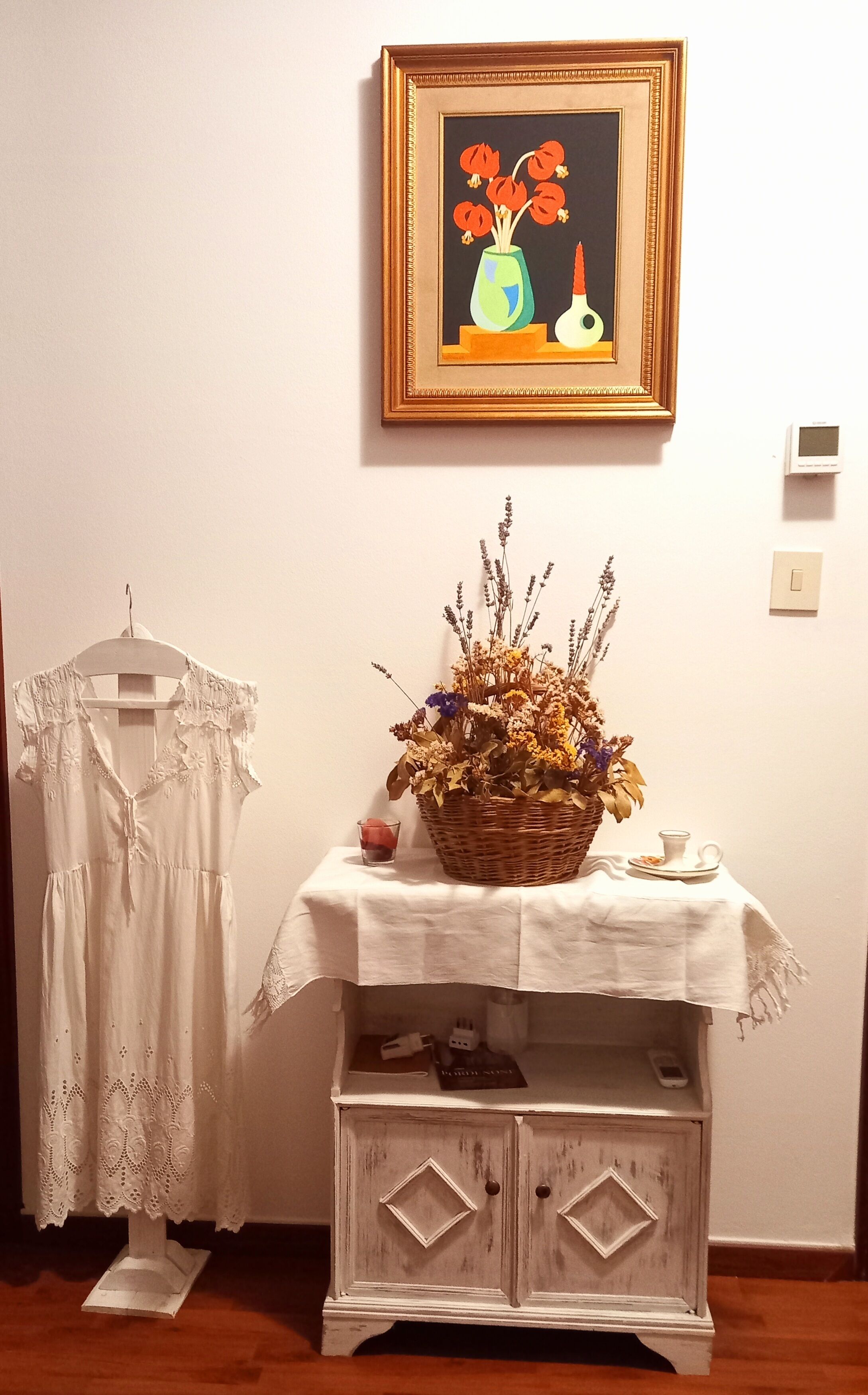 A room with a white dress on a hanger, a table with a basket of flowers, and a framed painting on the wall.