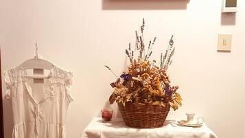 A room with a white dress on a hanger, a table with a basket of flowers, and a framed painting on the wall.