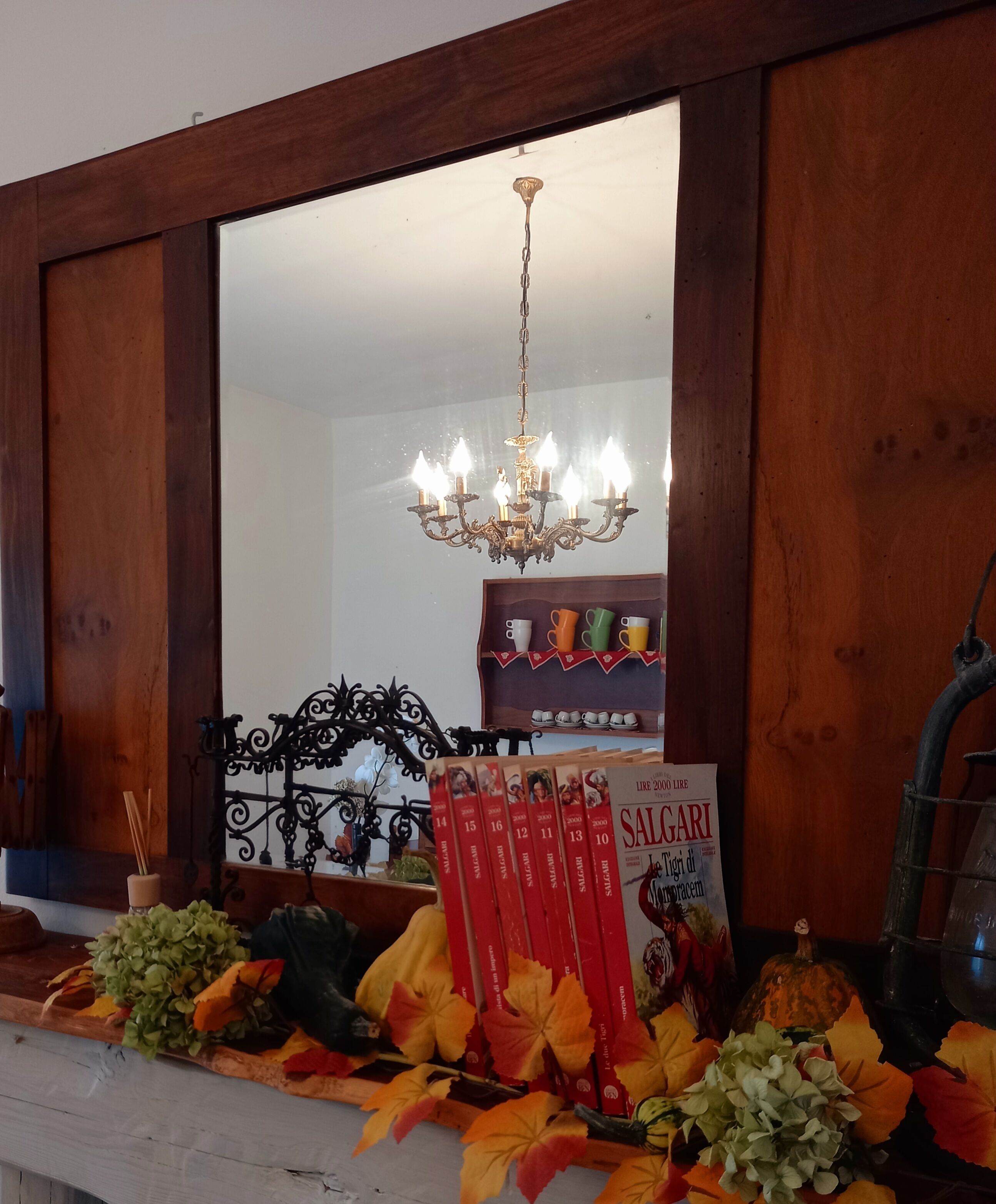 Uno specchio con cornice in legno, un lampadario sopra, una mensola con libri e un tavolo con foglie autunnali e una zucca.
