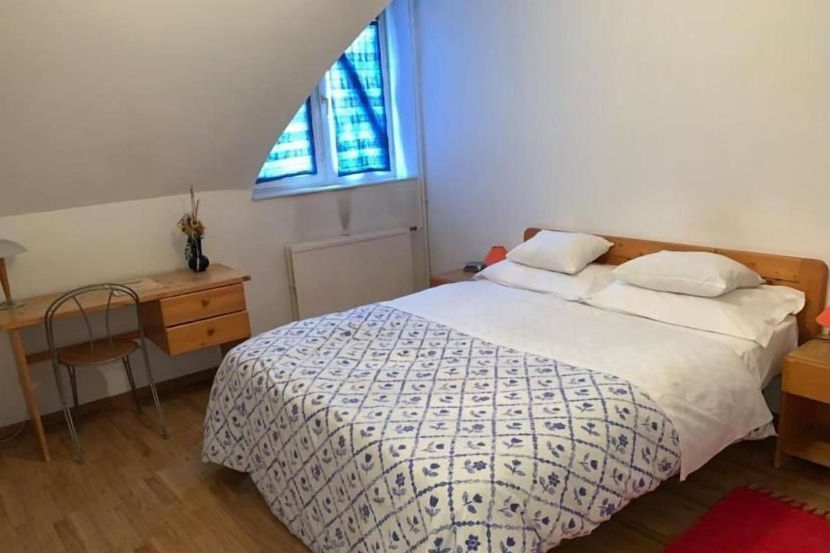 A bedroom with a bed, a desk, a chair, a window, and a radiator.