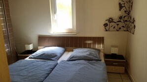 A room with two beds, a window, a wall-mounted artwork, and a nightstand with a lamp.