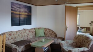 A cozy room with a sofa, a small table, and a chair. There is a painting on the wall and a door leading to another room.