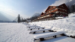 Una terraza de madera cubierta de nieve que conduce a un edificio de estilo chalete con varios balcones, con montañas al fondo.