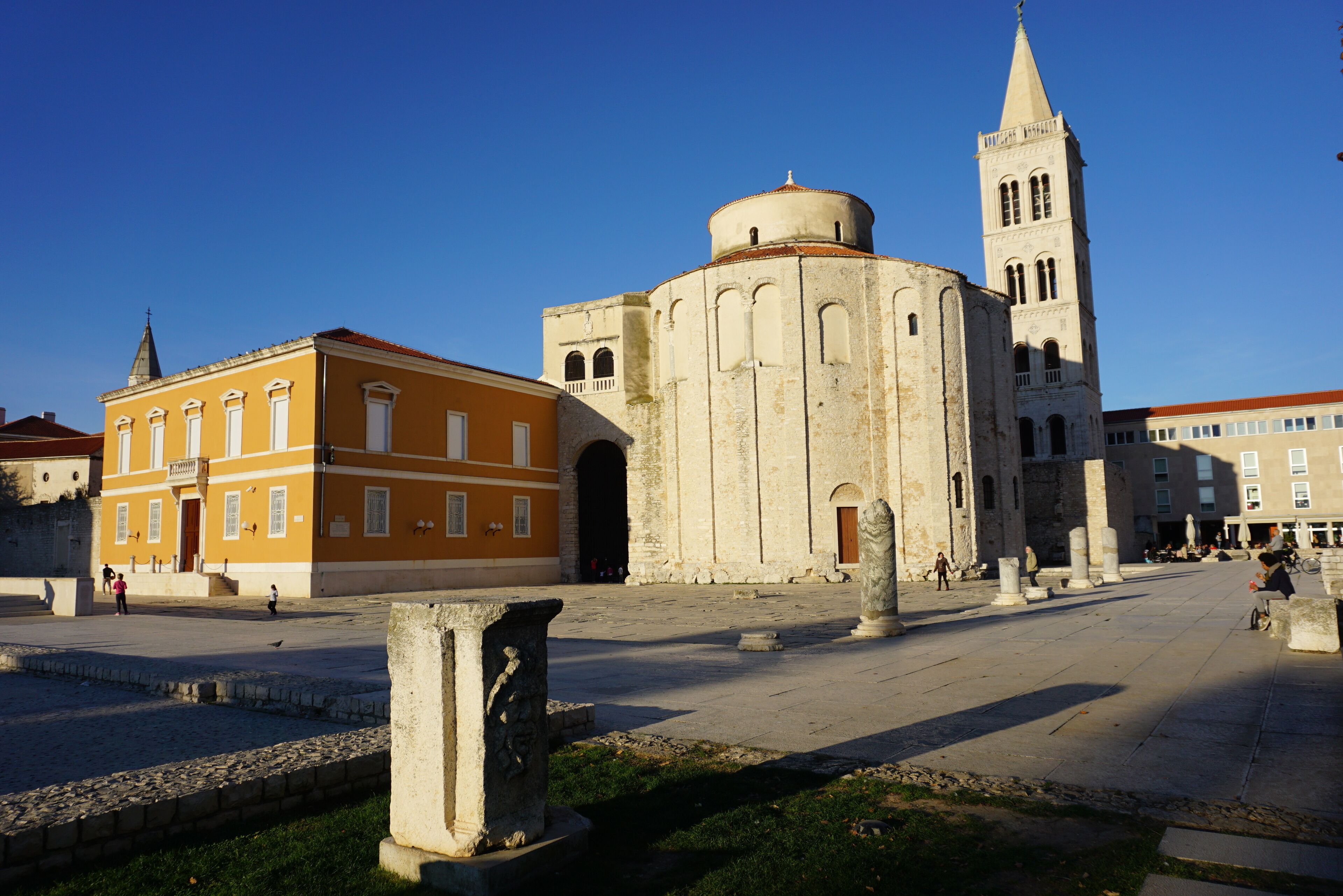 Une place historique, dominée par une église remarquable, dotée d’un clocher et bordée d’un bâtiment jaune aux fenêtres en arc.