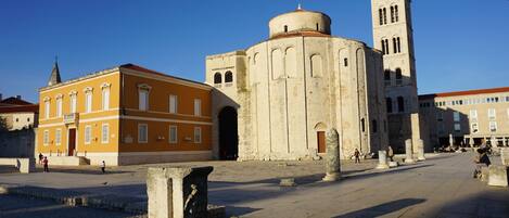 Une place historique, dominée par une église remarquable, dotée d’un clocher et bordée d’un bâtiment jaune aux fenêtres en arc.