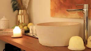 Ein Badezimmer mit einem runden, hellen Waschbecken, einem chromfarbenen Wasserhahn, einer brennenden Kerze und Dekorationselementen auf der Ablagefläche.