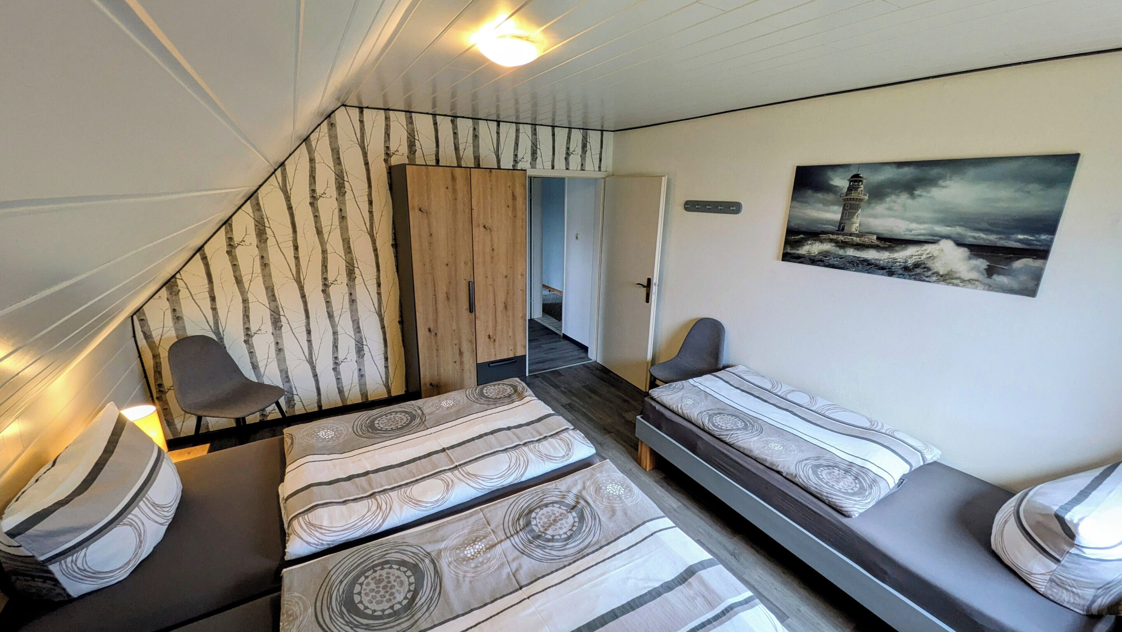 A compact room with two single beds, a wooden wardrobe, a chair, and a painting of a lighthouse.