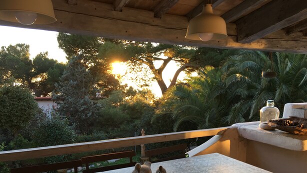 A balcony with a view of trees and a sunset. There is a table with a vase and a bottle.
