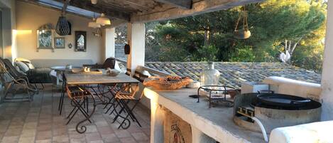 Um pátio externo com piso de ladrilho de pedra, pérgola de madeira, mesa com cadeiras e área de churrasco.