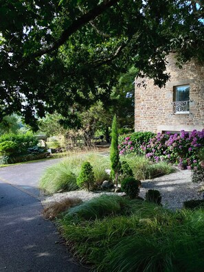 Un bâtiment en pierre avec une allée en gravier et un petit jardin agrémenté de fleurs violettes.