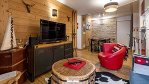 A cozy living room with a wooden wall, a mounted TV, a red leather sofa, and a dining area with a stone wall backdrop. - COZY APARTMENT RENOVATED CITY CENTER AVENUE LECLERC WIFI AND PRIVATE COVERED PARKING (CAUTERETTS)