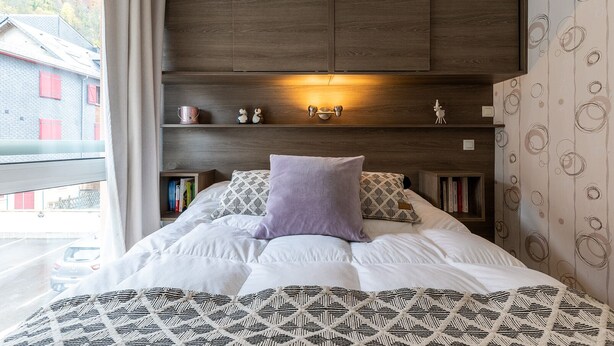 A bedroom with a bed, patterned bedding, a headboard with built-in lighting, a nightstand with books, and a window with a view of buildings. - COZY APARTMENT RENOVATED CITY CENTER AVENUE LECLERC WIFI AND PRIVATE COVERED PARKING (CAUTERETTS)